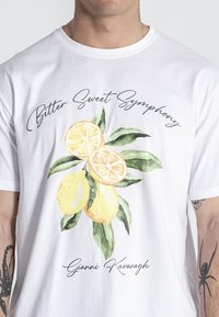 Biela bavlnená tričko so živou grafikou citrónu a listu, s textom "Bitter Sweet Symphony" a "Gianni Kavanagh" v elegantnom písmene.
