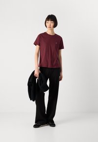 Woman standing against white background wearing a burgundy t-shirt, black pants, black shoes, holding a black knitted sweater in one hand.