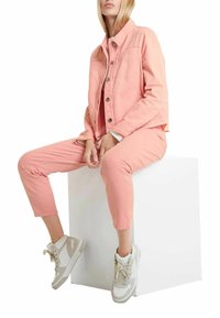 Kvinde sidder på hvid kube iført en ferskenfarvet denimjakke, matchende bukser og hvide høj-top sneakers med beige detaljer.