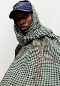 Rohelise ja musta hunditõmmisega sall pruunide narmastega äärtega ning tikandiga tekstiga, kantud ümber keha, koos mereväe nokamütsiga.