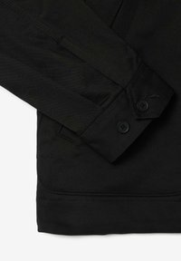 Melna jaka ar gludu auduma tekstūru, norādītu apkakli un divām melnām pogām pie piedurknes. Ietver tīrās, pielāgotās šuvju detaļas.