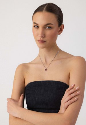 Junge Frau mit zurückgekämmtem Haar, trägt ein trägerloses schwarzes Oberteil und eine silberne Halskette mit einem herzförmigen Anhänger, die Arme verschränkt.