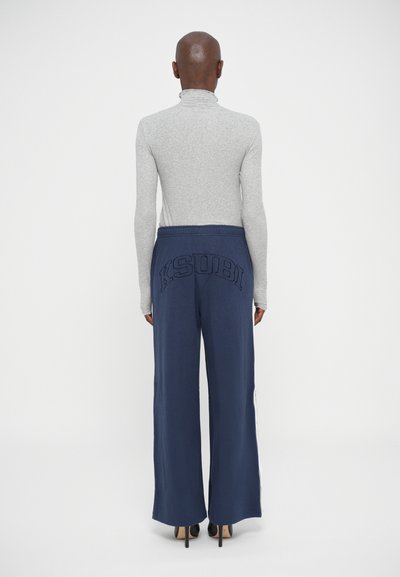 Persoon gekleed in marineblauwe wijde broek met het KSUBI-logo op de achterkant en een grijze lange mouwen coltrui, staand met het gezicht naar een witte achtergrond.