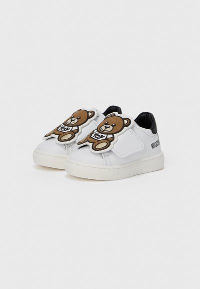 Λευκά συνθετικά παπούτσια με μαύρες λεπτομέρειες και δύο μεγάλα καφέ μπαλώματα αρκούδας στο μπροστινό μέρος. Στρογγυλερή μύτη και υφή στη σόλα.