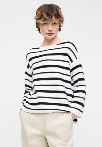 Νεαρό άτομο με κοντά καστανά μαλλιά φοράει λευκό πουκάμισο με μακριά μανίκια και μαύρες οριζόντιες ρίγες και μπεζ παντελόνι, τα χέρια στις τσέπες.