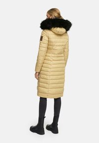 Beige polstret jakke med en sort faux fur hætte, lodret quiltning og en tætsiddende silhouette. Bæret med mørke jeans og sorte ankelstøvler.