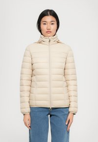 Beige gewatteerde jas met een genopt structuur, hoge kraag, volledige rits en elastische manchetten. Gedragen boven lichtblauwe jeans.