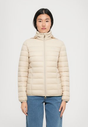 Beige gewatteerde jas met een genopt structuur, hoge kraag, volledige rits en elastische manchetten. Gedragen boven lichtblauwe jeans.