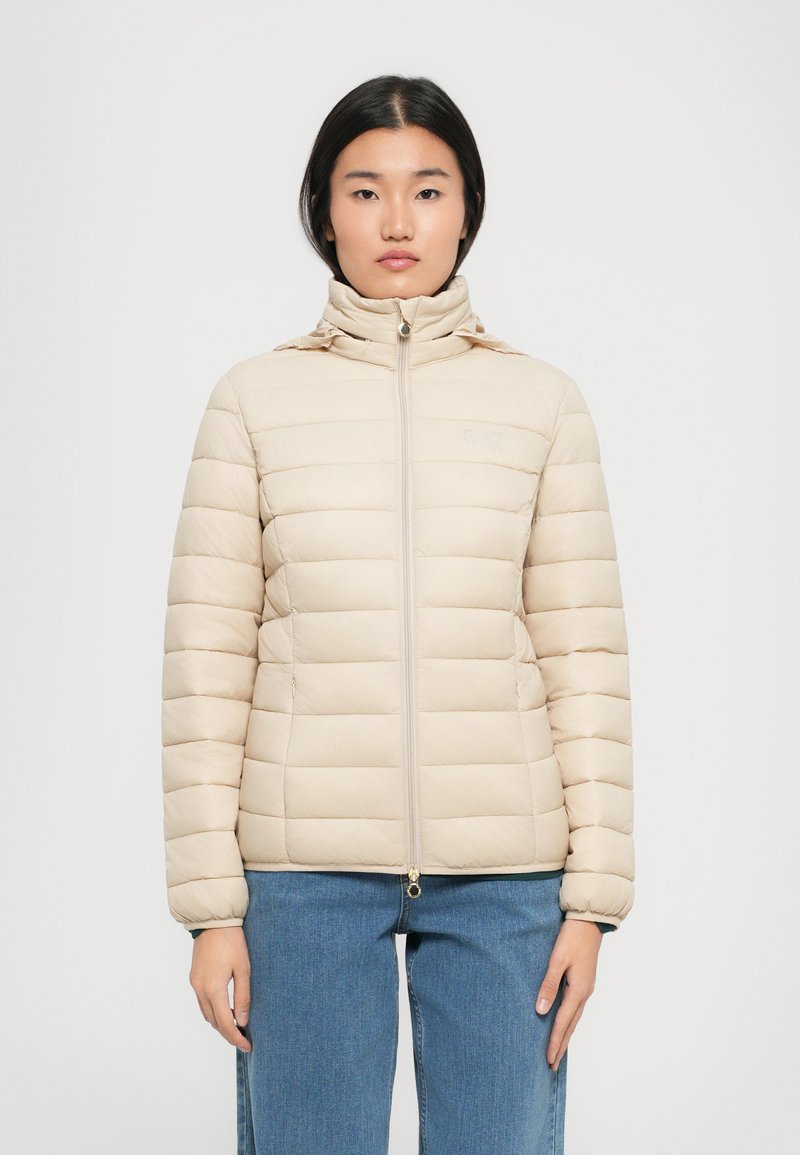 Beige gewatteerde jas met een genopt structuur, hoge kraag, volledige rits en elastische manchetten. Gedragen boven lichtblauwe jeans.