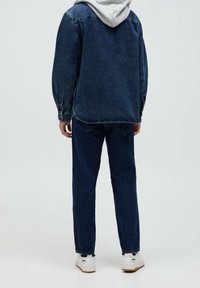 Person med en mörkblå jeansjacka och jeans samt en grå hoodie under, står vänd bort, vita sneakers synliga.