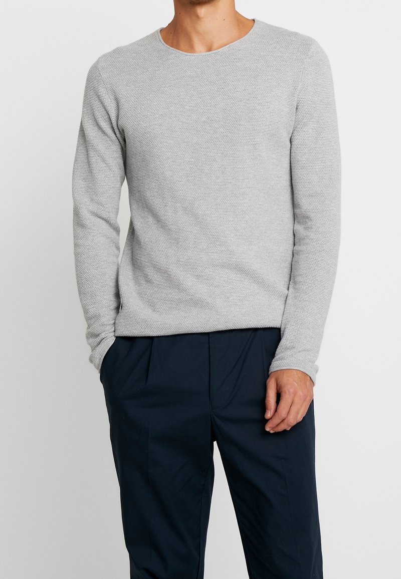 Homme portant un pull en maille à manches longues gris clair texturé avec un pantalon bleu marine, debout avec une main dans la poche sur un fond uni.