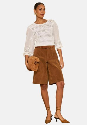 Nő fehér csipkefelsőt visel könyékig érő, puffos ujjakkal, barna színű, öves, térdig érő, szarvasbőr shortot, barna hátpántos magassarkút, kezében összecsukott barna dzsekit tart.