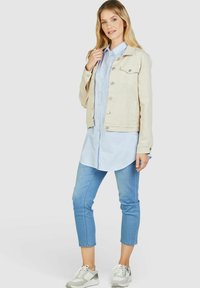 Hellbeige Jeansjacke mit Knopfverschluss, zwei Fronttaschen und tailliertem Design, kombiniert mit einem blau-weißen gestreiften Hemd und hellen Jeans.