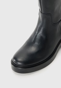 Gros plan sur l'avant et le côté d'une botte en cuir noir avec un embout arrondi et une semelle épaisse sur un fond clair.