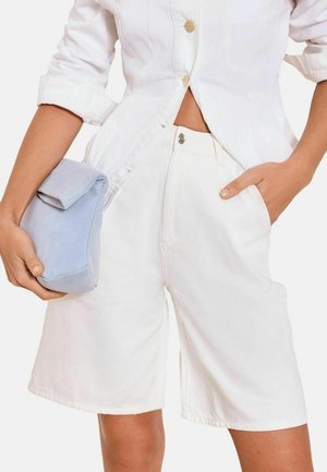 Kvinde iført hvid knappet bluse og højtaljede hvide shorts, der holder en lille lyseblå ruskindspung i den ene hånd.