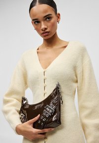 Γυναίκα με μαλλιά χτενισμένα προς τα πίσω, φορώντας ένα κρεμ πλεκτό ζακέτα, κρατά μία τσάντα από σκούρο καφέ δέρμα με ασημένια αγκράφα και λεπτομέρειες αλυσίδας.