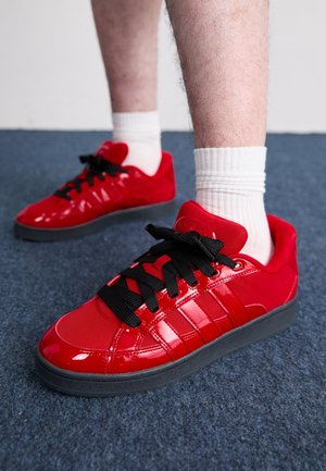 Personne portant des baskets rouges brillantes avec des lacets noirs et des chaussettes blanches montantes debout sur un tapis bleu foncé.