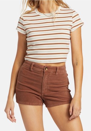 Vrouw draagt een wit crop top met bruine strepen en hoogwater bruine corduroy shorts, staand met ontspannen handen langs het lichaam.