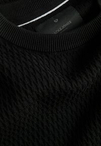 Pull noir tricoté avec un col côtelé et un motif torsadé texturé visible, avec une étiquette Jack & Jones partiellement visible à l'intérieur du col.