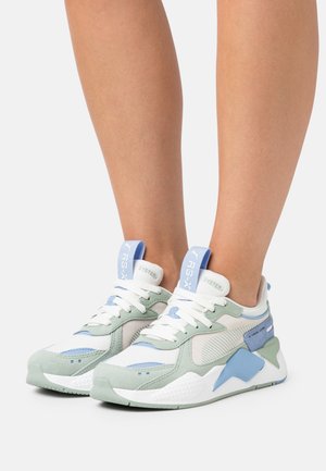 Personne portant des baskets blanches, vert clair et bleues avec des lacets, debout sur une surface blanche devant un fond uni.