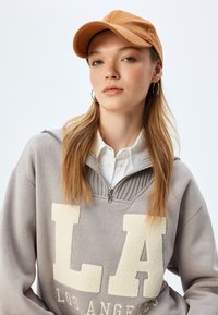 Szary sweatshirt z białym napisem "LA", z suwakiem na pół długości, nałożony na białą koszulę z kołnierzykiem. Brączowa czapka o miękkiej fakturze.