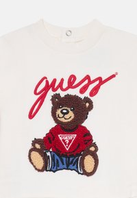 Balta šviesiai mėlyna su rausvu lokiu, siuvinėtu raudoną megztinį, featuring "Guess" logotipą ir mėlynas šortus. Apvalaus kaklo dizainas.