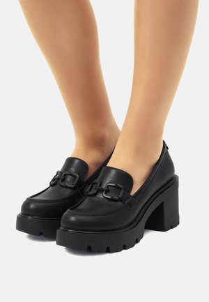 Zapatos de plataforma de cuero negro con un tacón grueso, que presentan una textura suave, costuras prominentes y un detalle de metal en la parte delantera.