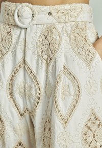 Pantaloni in lino con intricati ricami beige con motivi floreali e paisley. Vita alta con passante rotondo per la cintura e dettaglio con bottone.