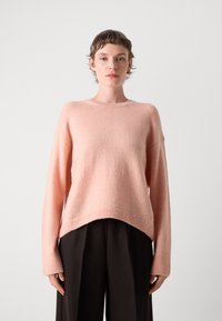 Pull en laine mélangée rose avec une coupe ample, des épaules tombantes et un ourlet côtelé. Associé à un pantalon foncé, présentant une texture douce et lisse.
