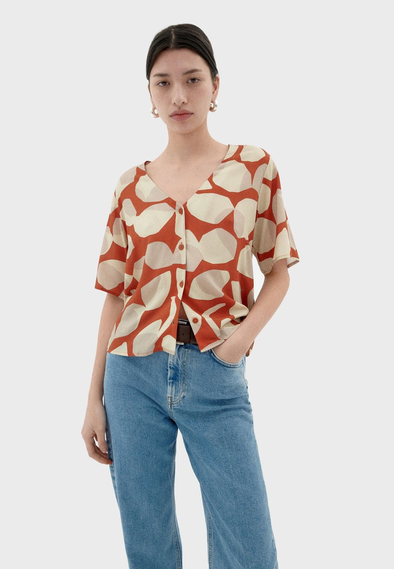 Kurzärmliges Hemd in Creme mit einem auffälligen roten abstrakten Muster. Hat einen V-Ausschnitt und eine lockere Passform. Kombiniert mit blauen Jeans.