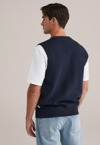 Mornarice plava pletena prsluk s okruglim izrezom, nosi se preko bijele majice. Ima rebraste rubove i malu oznaku s logom na rubu.