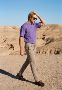 Jeune homme portant des lunettes de soleil, un t-shirt violet, un pantalon beige et des chaussures marron marchant sur un paysage désertique sec et rocheux sous un ciel bleu clair.
