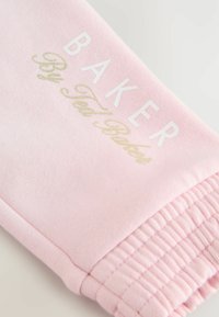 Sudadera rosa claro con puños acanalados y texto de logo impreso en blanco y dorado, fabricada con un tejido suave y texturizado.