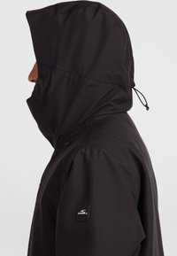 Veste imperméable noire avec un col haut et une capuche, dotée de cordons de serrage réglables et d'un patch de marque sur la manche gauche.