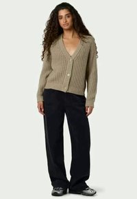 Beige stickad cardigan med djup V-ringning, stora knappar och strukturerat mönster. Matchad med lösa svarta byxor och casual skor.