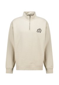 Beige pullover sweatshirt with a quarter zip, ribbed cuffs, and a small black graphic design on the left chest. Soft cotton texture.