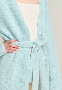 Light blue knitted cardigan with a textured finish, open front, and a tied belt detail at the waist. Soft, cozy fabric is featured.
