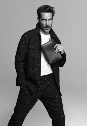 Black textured blazer and pants with white flecks; white t-shirt; black leather clutch held in hand; model poses confidently.