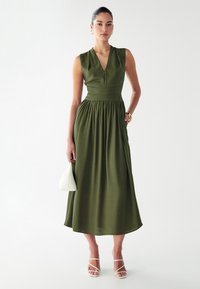 Vestido sin mangas de color verde oliva con escote en V, cintura ceñida y falda fluida. La modelo sostiene un bolso blanco y lleva sandalias blancas con tiras.