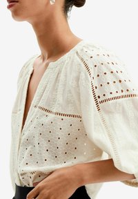 Blusa bianca con motivo floreale ricamato, scollo aperto e maniche a palloncino con dettagli a taglio lungo le cuciture. Texture morbida.