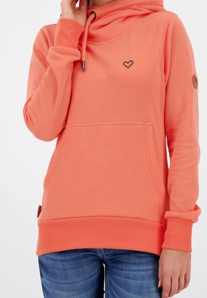 Personne portant un sweat à capuche corail avec poche avant et emblème en forme de cœur, associé à un jean bleu, sur un fond clair uni.