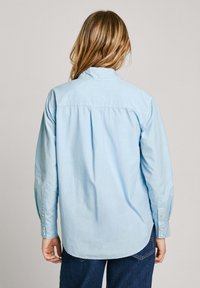 Camicia azzurra a maniche lunghe con colletto a bottoni, orlo arrotondato e piega sul retro; tessuto morbido e leggero; polsini arrotolati con bottoni.
