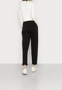 Person som bär svarta raka byxor, vit långärmad topp och vita Veja-sneakers, står med händerna i fickorna mot en enkel vägg.