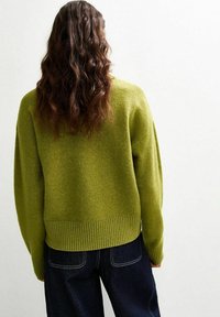 Pull en maille vert, à la coupe ample, avec un ourlet côtelé et de larges manches, associé à un jean en denim foncé. Texture douce visible.