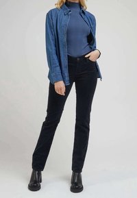 Camisa de mezclilla azul de botones sobre un cuello de tortuga azul con canalé, combinada con pantalones de pana negros y botines de tacón negros.