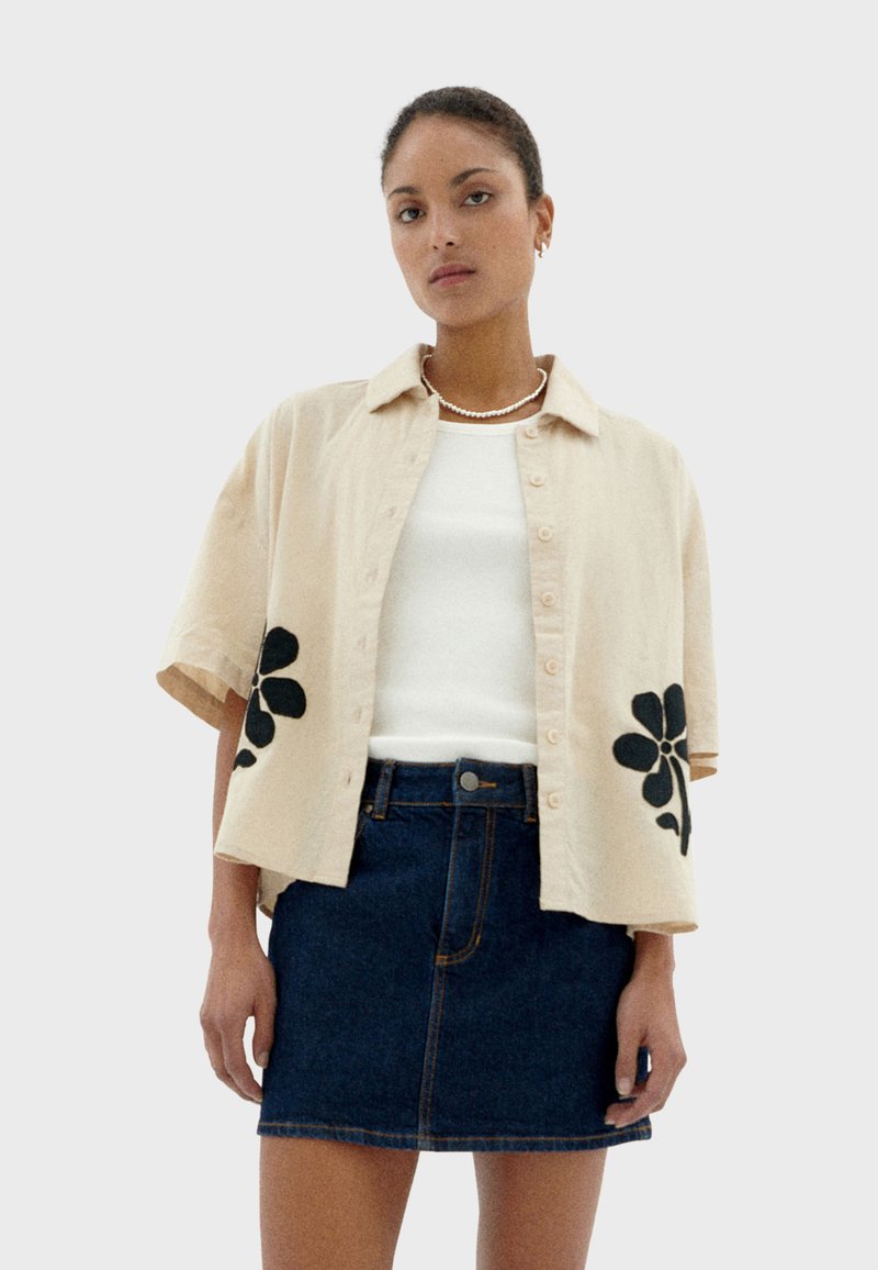 Beige short-sleeve shirt with black floral embroidery on hem, worn over a white top, paired with a dark denim mini skirt.