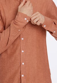 Camicetta a maniche lunghe color ruggine realizzata in tessuto texturizzato, con bottoni bianchi, chiusura con bottoni frontale e polsini regolabili.