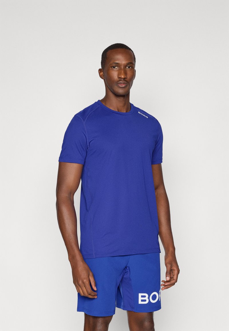 Niebieska koszulka sportowa z krótkimi rękawami i okrągłym dekoltem, w połączeniu z dopasowanymi niebieskimi szortami z białym napisem "BORG". Gładka tekstura.