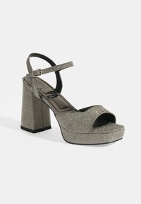 Hoge hak sandaal in metallic zilveren glitter. Beschikt over een open neus, enkelbandje en een chunky blockhak met een platformzool.