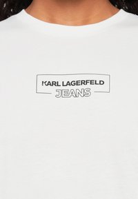 Balta kokvilnas t-kreklā ar melnu izdrukātu logo, kas lasāms "KARL LAGERFELD JEANS." Īsas piedurknes un klasiskā apaļā kakla izgriezums.